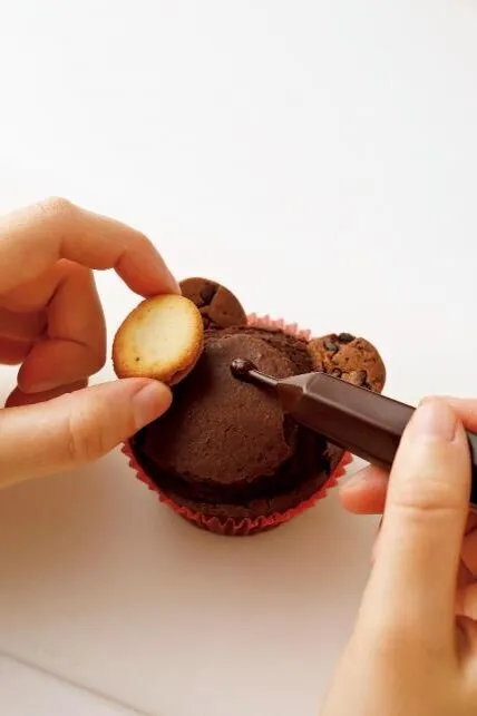 チョコペンを湯につけて柔らかくし、顔の中央に少量絞って、口もと用のクッキーをはりつける。チョコペンで目、鼻、口を描く。

（画像：口もと用のクッキーをはりつける。）