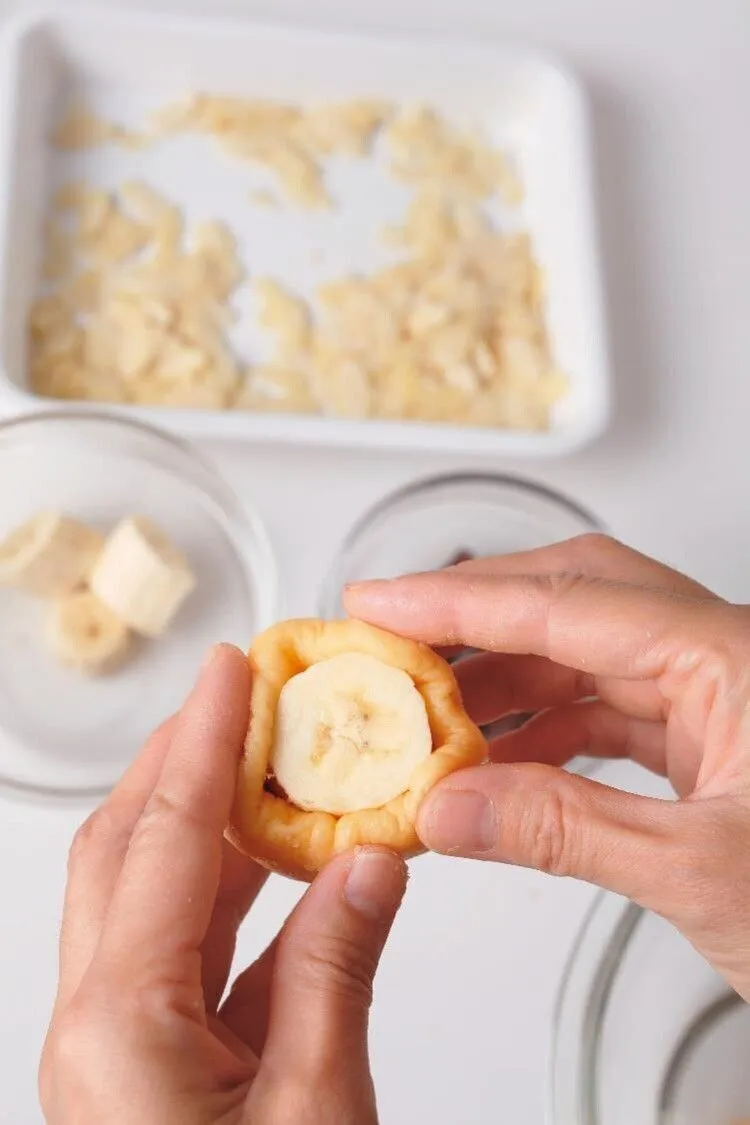 生地を6等分にして直径5cmくらいの円形にのばし、チョコの1/6量、バナナ1切れを真ん中にのせる。生地の縁を少しずつ持ち上げるようにして包み、包み終わりをしっかりと閉じて丸く形を整える。まわりに、アーモンドスライスをまんべんなくつける。残りも同様に作る。

（画像：生地でチョコ、バナナを包む。）