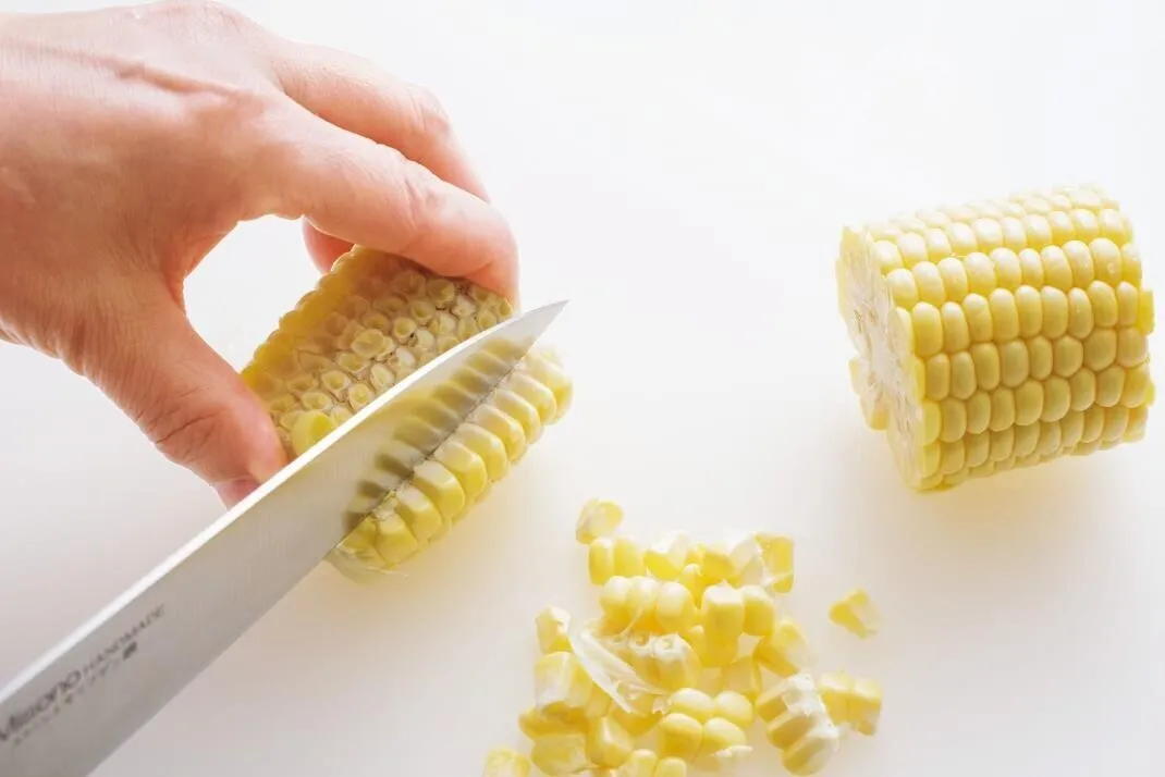 とうもろこしは皮とひげを取って長さを3等分に切り、身を切り落とす。玉ねぎはみじん切りにする。トマトはへたを取り、横半分に切って種を取り、1cm角に切る。エビは背わたがあれば取って洗い、水けを拭く。

（画像：とうもろこしは身を切り落とす。）