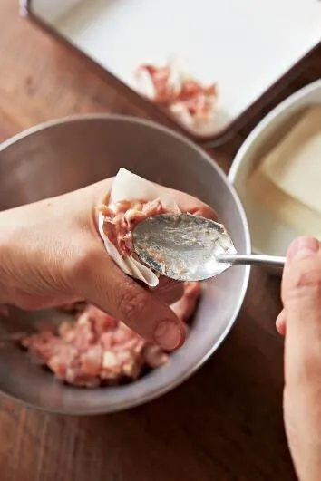 ボールにしいたけ以外のAのたねの材料を入れ、粘りが出るまで手で練り混ぜる。しいたけを加えてさらに混ぜる。親指と人さし指で輪を作ってシュウマイの皮1枚をのせ、輪の中にスプーンでたねの1/16量を押し込むようにして入れ、円筒形に整える。残りも同様にする。

（画像：皮にたねを詰める。）