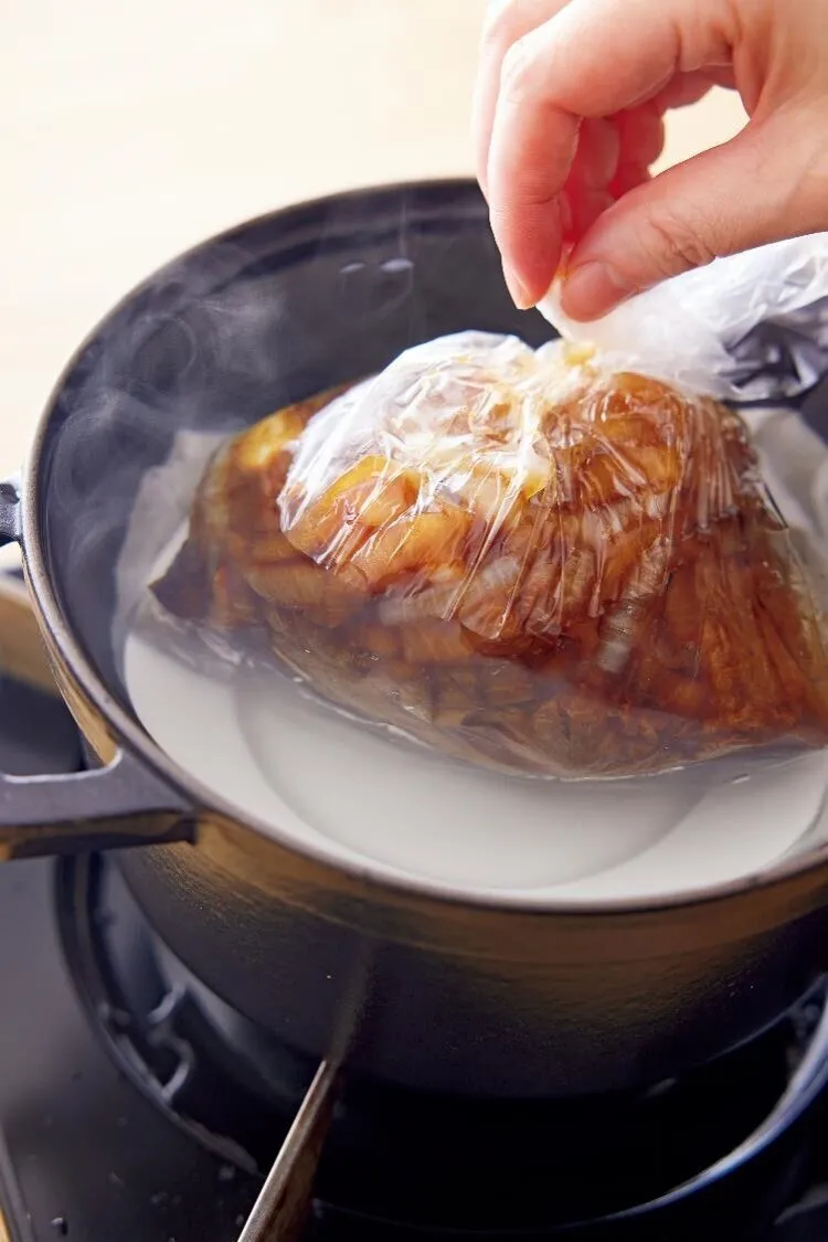 口径20cmの鍋にふきん※、耐熱の小皿を順に入れ、たっぷりの湯（1.2lが目安）を沸かす。耐熱のポリ袋（耐熱温度120℃以上の食品用ポリ袋を必ず使用してください（「アイラップ」＜岩谷マテリアル＞など）。耐熱性が低いと加熱中に溶けるおそれがあります。）に牛肉、フライパンの具とたれをすべて入れ、空気を抜いて口を閉じる。牛肉をポリ袋ごと耐熱の小皿の上にのせ、ふたをして火を止め、そのまま30分おく。
※沸騰の振動で器が割れるのを防ぐため、鍋底に敷く。

（画像：ポリ袋ごと小皿の上にのせる。）